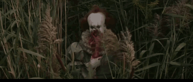 a clown is standing in a field of tall grass holding a bloody axe .