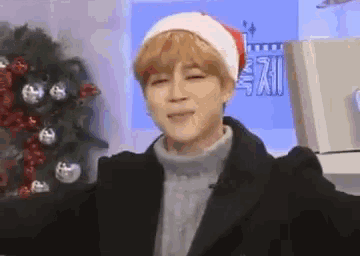 a young man wearing a santa hat and a sweater is standing in front of a christmas tree .