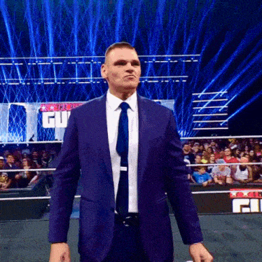 a man in a purple suit and tie is standing in a wrestling ring with a sign that says cu on it