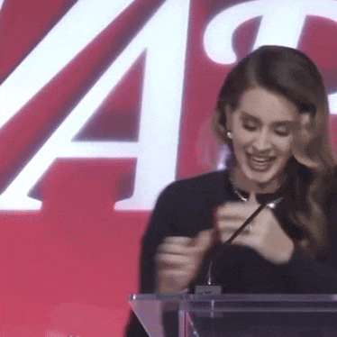 a woman is sitting at a podium with a microphone in front of a red wall .