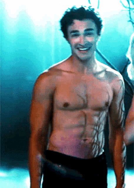 a shirtless man smiles while standing in front of a blue background