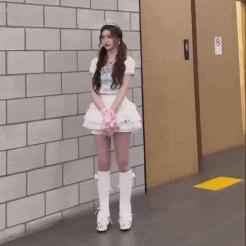 a girl in a white dress and pink gloves is standing in front of a brick wall