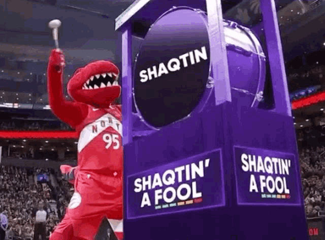a raptor mascot is holding a hammer in front of a large drum that says shaqtin ' a fool