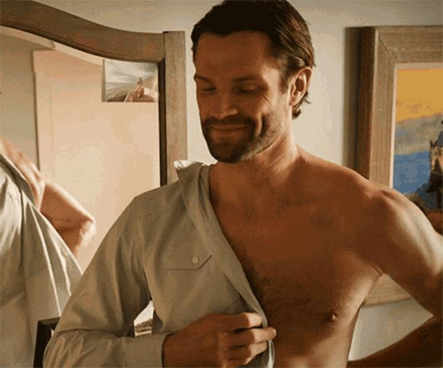 a shirtless man adjusts his shirt in front of a framed picture