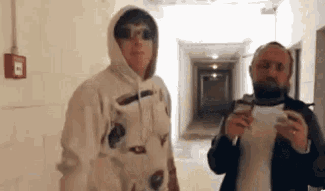 two men are standing next to each other in a hallway . one of the men is wearing a hoodie and sunglasses .
