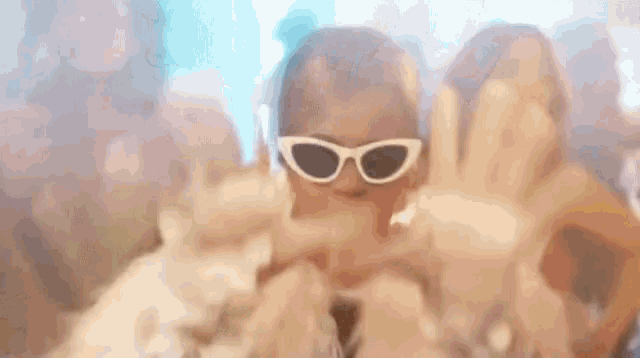 a woman wearing sunglasses is making a peace sign with her hands in front of a crowd of people .