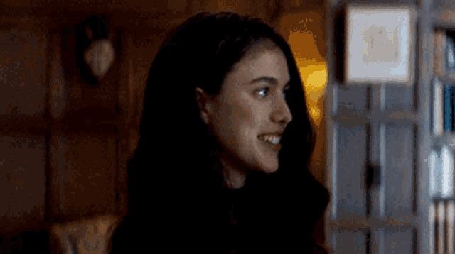 a woman with long black hair is smiling in a dark room with a bookshelf in the background .