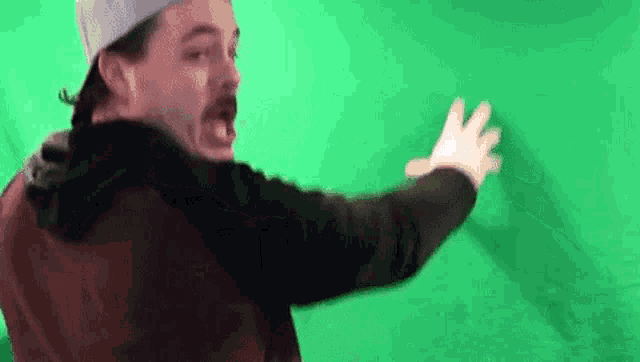 a man in a baseball cap is standing in front of a green screen and making a funny face .