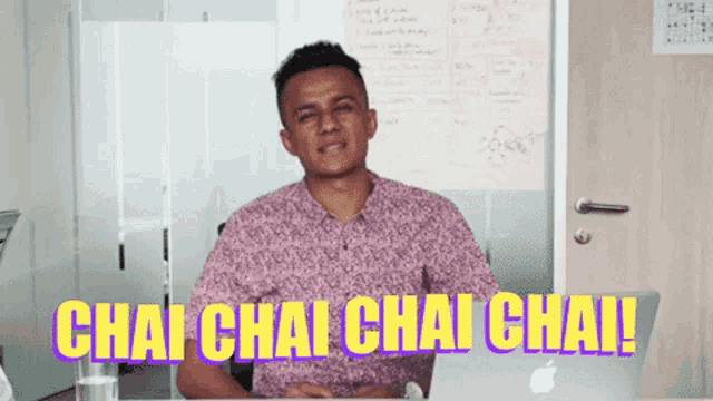 a man in a purple shirt is sitting at a desk with a laptop and says chai chai chai chai !