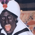 a man with black face paint is wearing a white turban and sticking his tongue out .