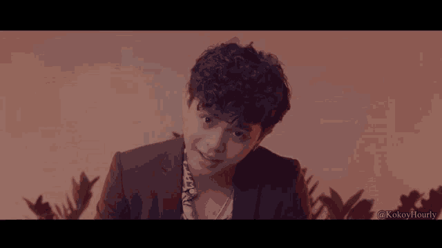 a young man with curly hair is smiling in front of a pink background with the hashtag @kokoyhourly at the bottom