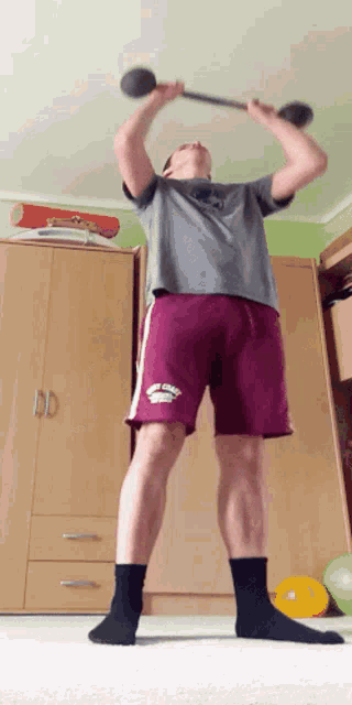 a man in a gray shirt and maroon shorts holds a barbell above his head