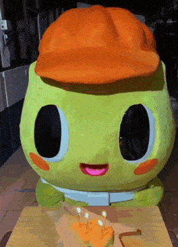 a green stuffed animal wearing an orange hat is sitting in front of a cake with candles and the number 7 on it