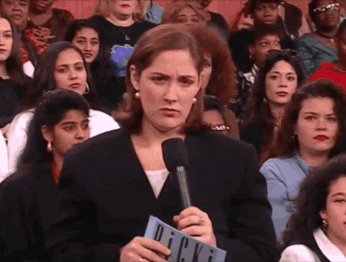 a woman stands in front of a crowd holding a microphone and a piece of paper with the word dick on it