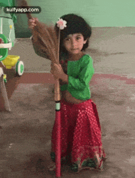 a little girl in a green shirt and red skirt is holding a broom and a flower .
