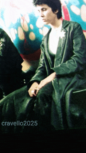 a woman in a green leather coat sits in front of a colorful background with the year 2025 on the bottom