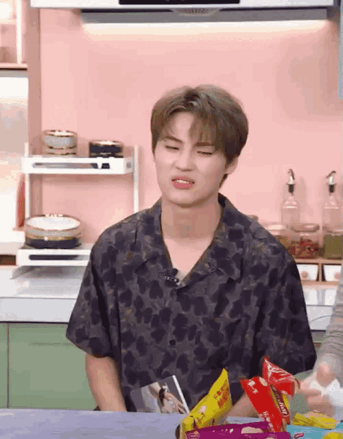 a young man in a floral shirt is sitting in a kitchen with a bag of chips in front of him