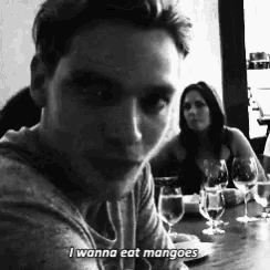 a man is sitting at a table with a woman behind him and says i wanna eat mangoes