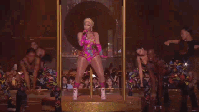 a woman in a pink bodysuit is standing on a pole in front of a group of dancers .