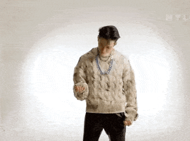 a man in a sweater and necklace is standing in front of a white wall .