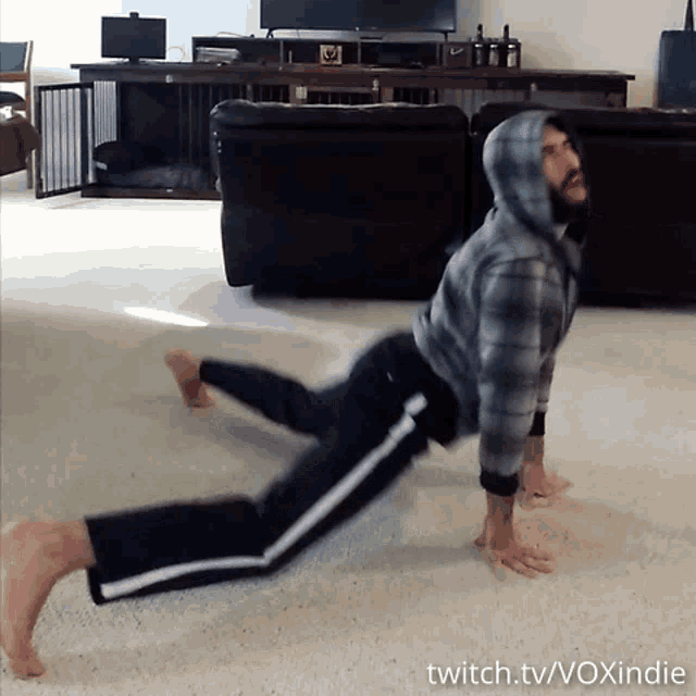 a man in a plaid hoodie stretches his legs in front of a couch with twitch.tv/voxindie written below him