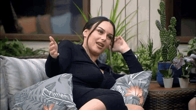 a woman in a black dress is sitting on a couch with pillows and a cactus in the background .