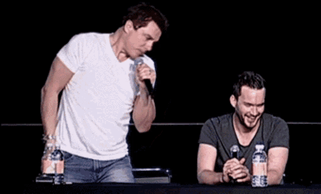 two men are sitting at a table with microphones and water bottles in front of them .