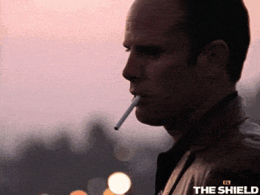 a man in a leather jacket is smoking a cigarette with the shield logo on the bottom