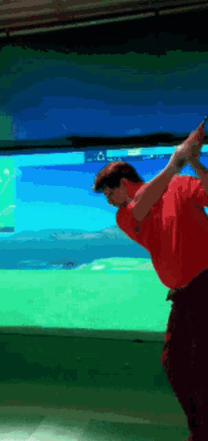 a man in a red shirt is swinging a golf club in front of a screen