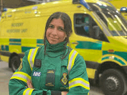 a female paramedic is standing in front of an ambulance with her arms crossed .