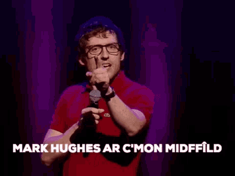 a man with glasses and a red shirt is holding a microphone and making a gesture with his finger .