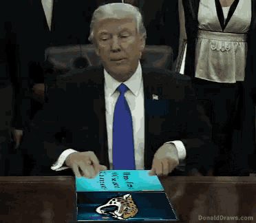 a man in a suit and tie is sitting at a table holding a book that says donald draws.com on it