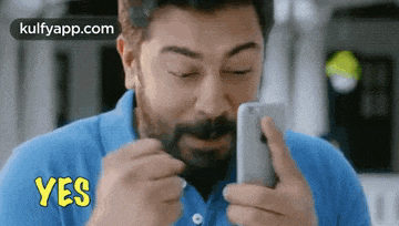a man in a blue shirt is holding a cell phone in his hand and says yes .