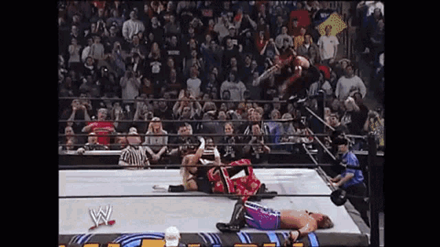 a wrestler is jumping in the air while another wrestler is laying on the ground in a wrestling ring .