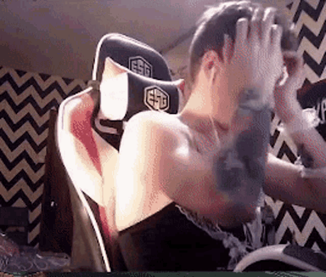 a man with a tattoo on his arm is sitting in a gaming chair covering his face .