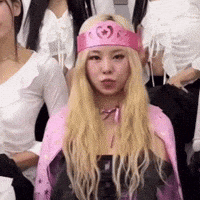 a blonde woman wearing a pink crown and a pink cape is standing in a crowd of people .
