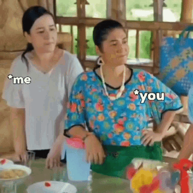 two women are sitting at a table with their hands on their hips . one of the women is wearing a floral shirt .