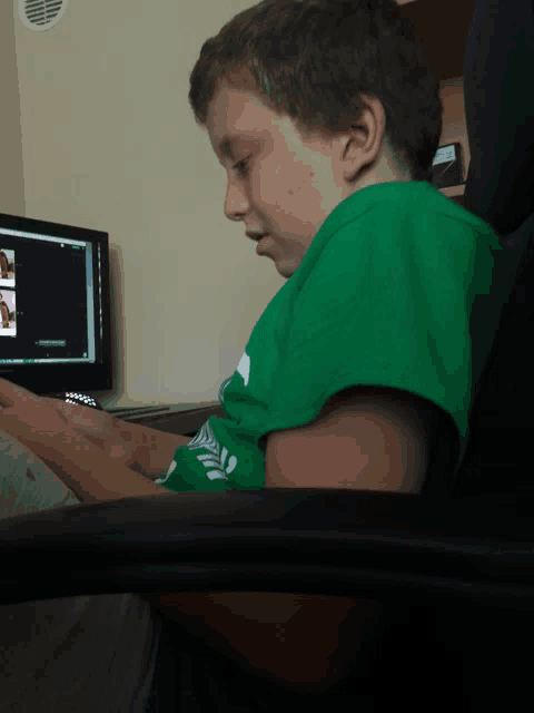 a young boy in a green shirt sits in front of a computer screen