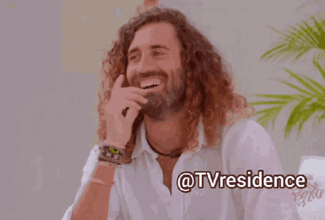 a man with long curly hair and a beard is smiling and holding his hand to his head ..