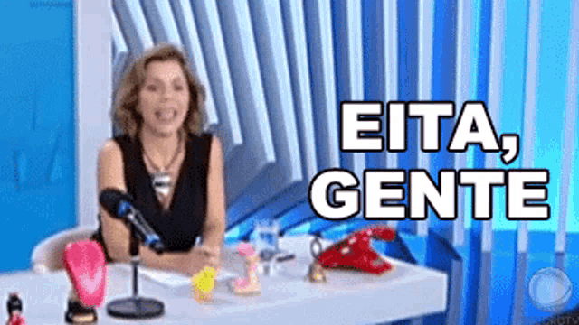 a woman is sitting at a desk in front of a microphone with the words " eita gente " written above her