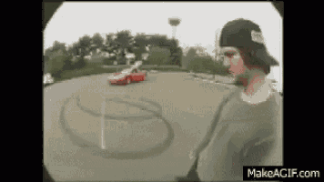 a man in a baseball cap is standing in front of a red car that is doing a stunt .