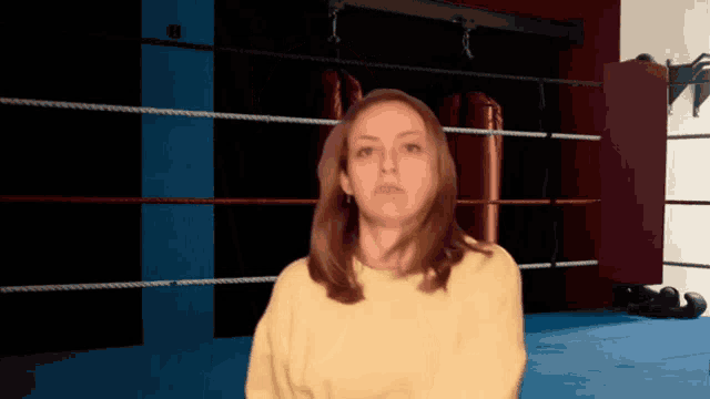 a woman in a yellow sweater is standing in a boxing ring looking at the camera