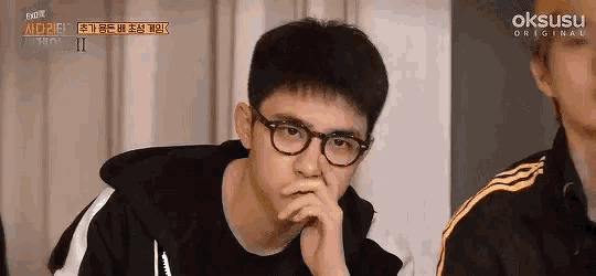a young man wearing glasses is sitting with his hands on his chin .