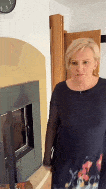 a woman in a black dress is standing in front of a fireplace with a clock on the wall