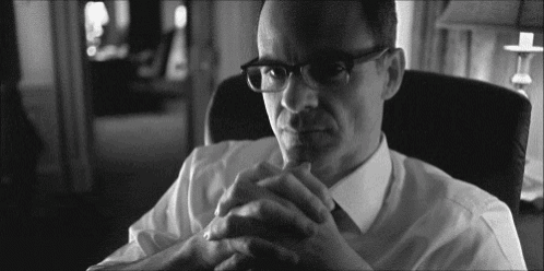 a man wearing glasses is sitting in a chair with his hands folded in front of him .