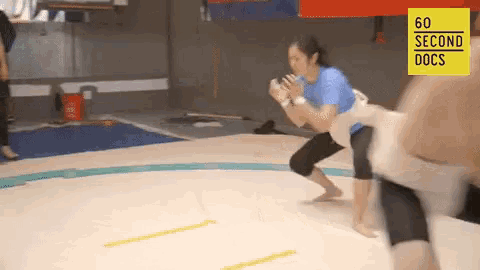 a woman in a blue shirt is standing on a wrestling mat with a yellow sign that says 60 second docs on it .