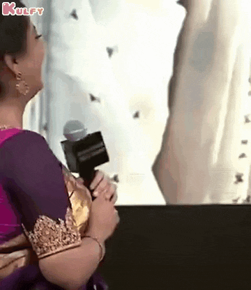 a woman in a purple saree is holding a microphone and talking into it .