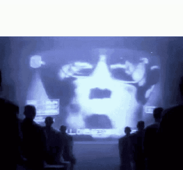 a group of people are standing in front of a large screen with a man 's face on it