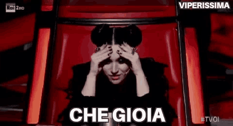 a woman is sitting in a red chair with her hands on her head and says che gioia .