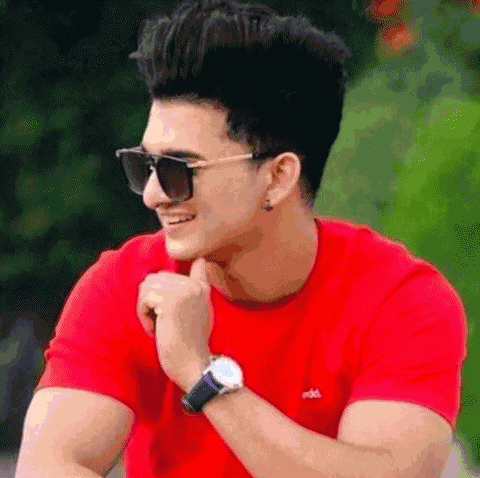 a man wearing sunglasses and a red t-shirt is smiling while holding his hand to his chin .